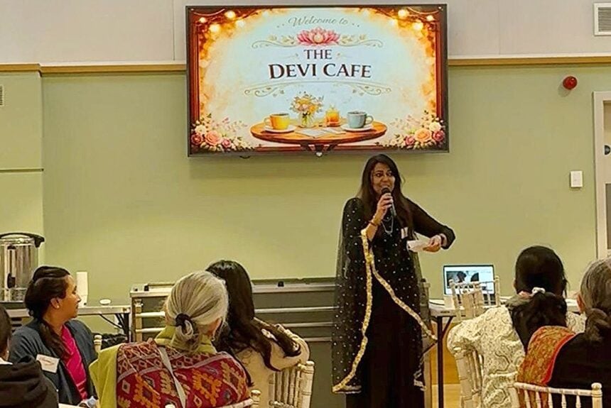 Speaker addresses attendees at The Devi Cafe, holding a microphone beneath a welcome screen with teacups; an event focused on empowering, protecting and inspiring women, and testing ideas.