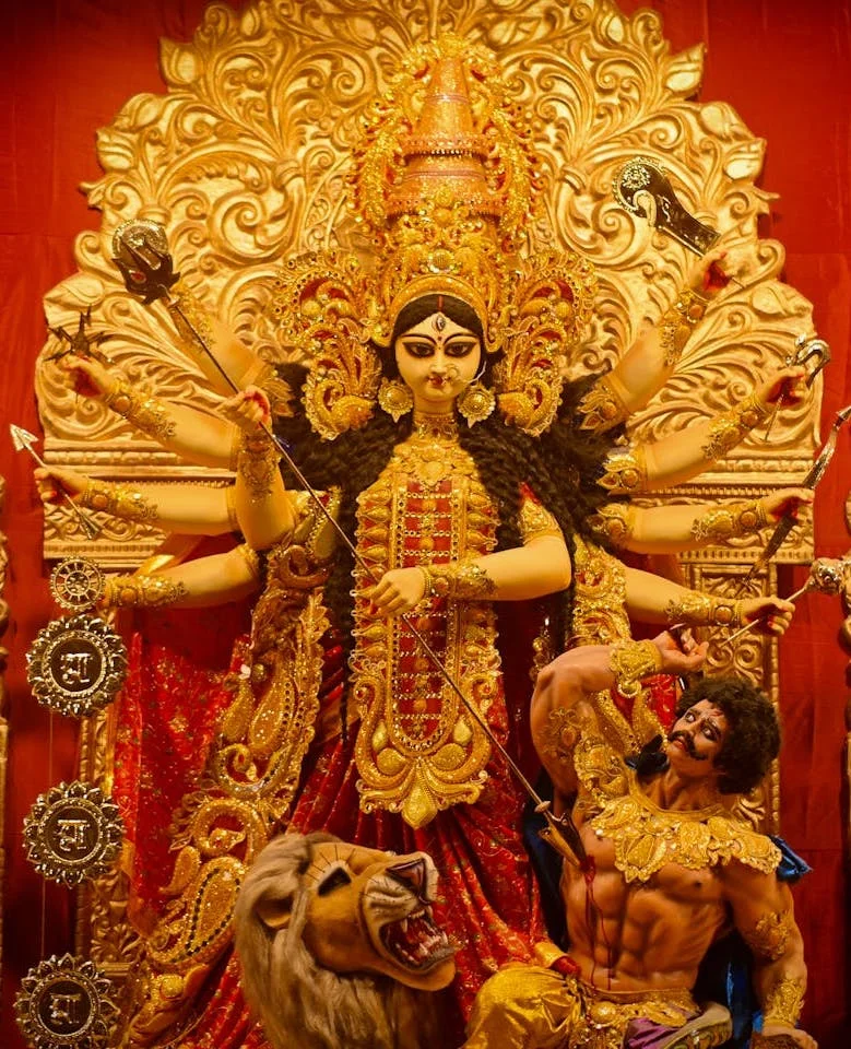 Golden idol of Goddess Durga in red sari, ten arms holding weapons, slaying Mahishasura beside a roaring lion, set against an ornate gold backdrop during the Navratri festival.