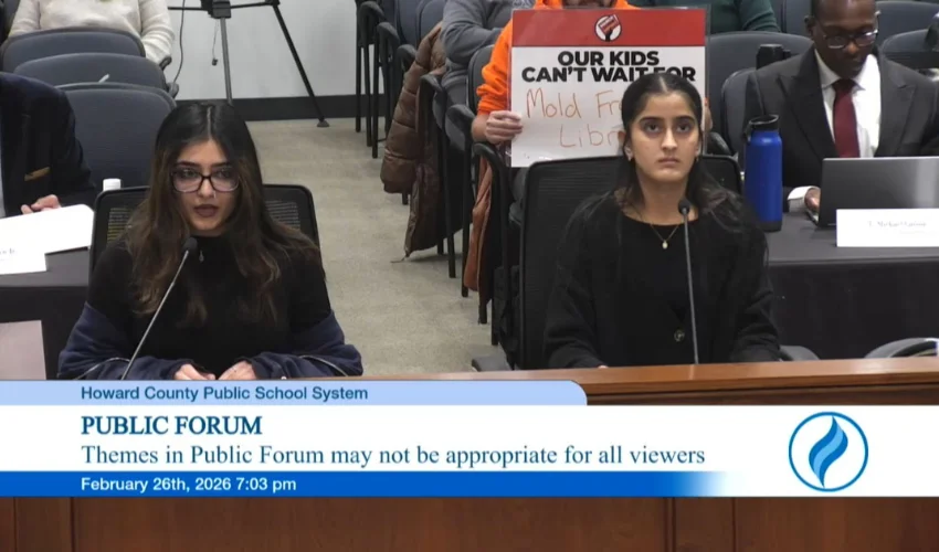 Two speakers address a Howard County Public School System board meeting, seated at microphones, as attendees watch; a sign behind reads 'Our Kids Can’t Wait—Mold Free Library.'