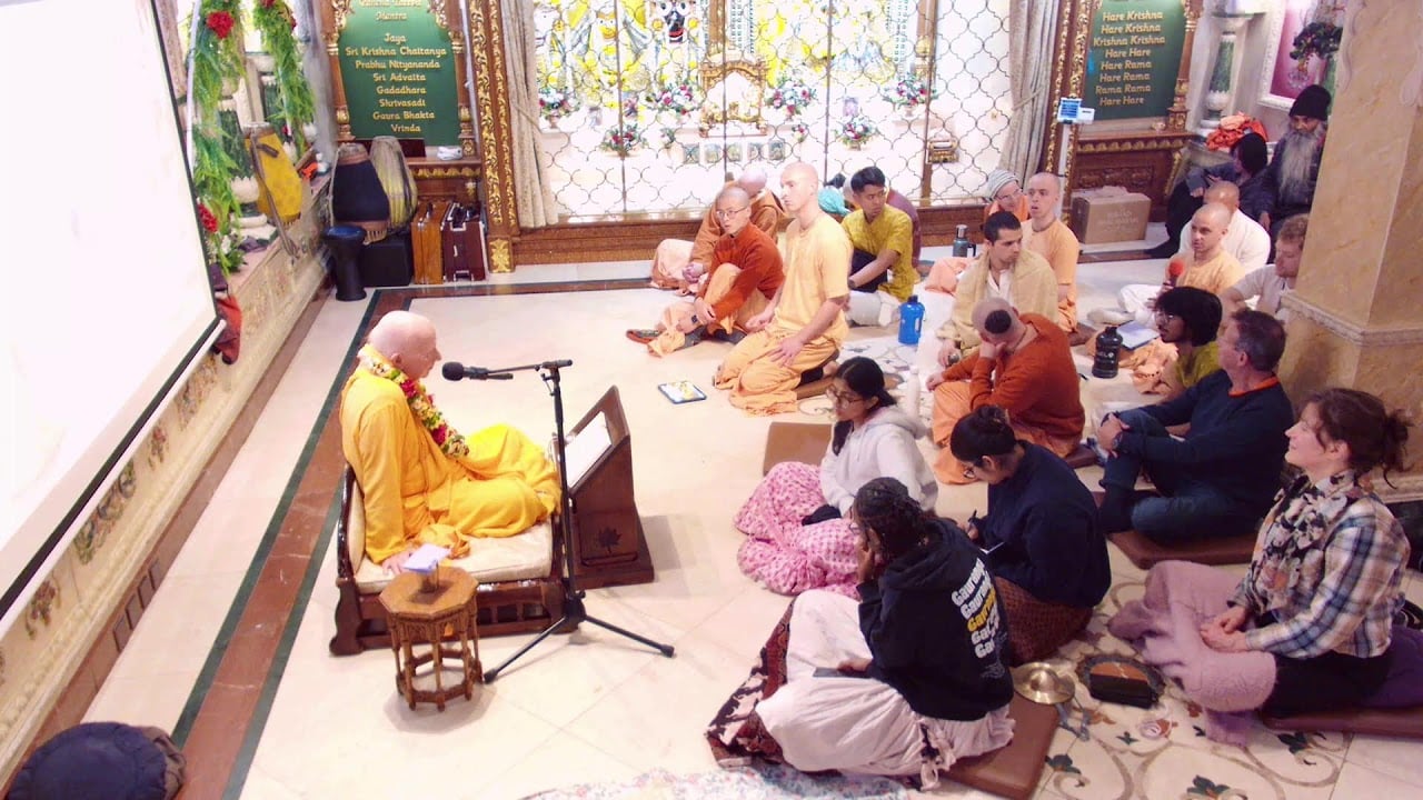 Temple lecture: a saffron-robed monk speaks into a mic from a low seat as devotees listen on the floor; ornate altar and mantra boards behind. SB 2.7.17 class, 01.04.2026, testing.