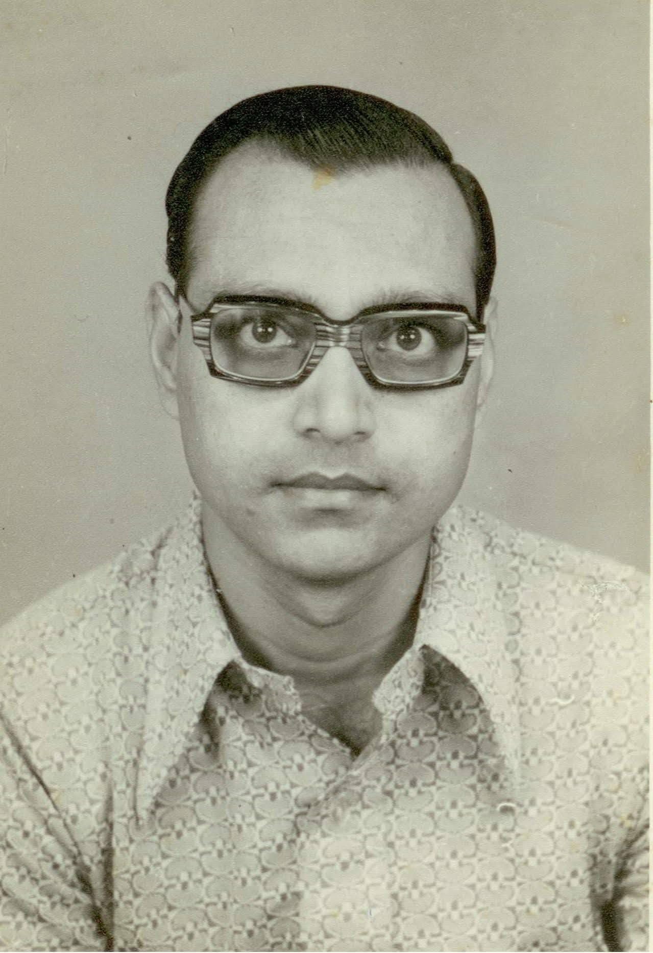 Monochrome studio portrait of a person facing the camera, wearing thick rectangular glasses and a patterned shirt on a plain backdrop; used with the feature 'The Blessing of Eons'.