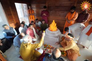 Priests and devotees perform puja in a wooden shrine around a white altar topped by an orange artificial flame, a stylized Akhand Jyoti symbol at a Ram temple, with flowers and ritual offerings.