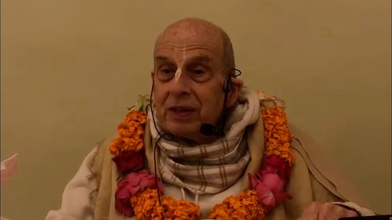 Speaker wearing a headset mic, shawl, and bright flower garlands sits before a plain wall during a Morning Class on SB 10.4.20, captured 17 April 2026 for testing and archive use.