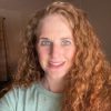 Close-up of a smiling adult with long curly red hair in warm indoor light, wearing a mint-green shirt and facing the camera. Image for parenting blog on breaking cycles, repair, generational healing.