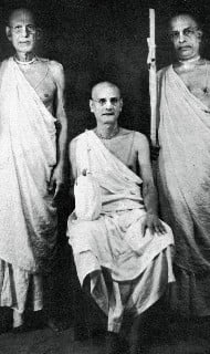 Black-and-white portrait of three elderly men in simple, draped garments; one seated at center, two standing beside him, all facing the camera against a plain backdrop, composed and dignified.