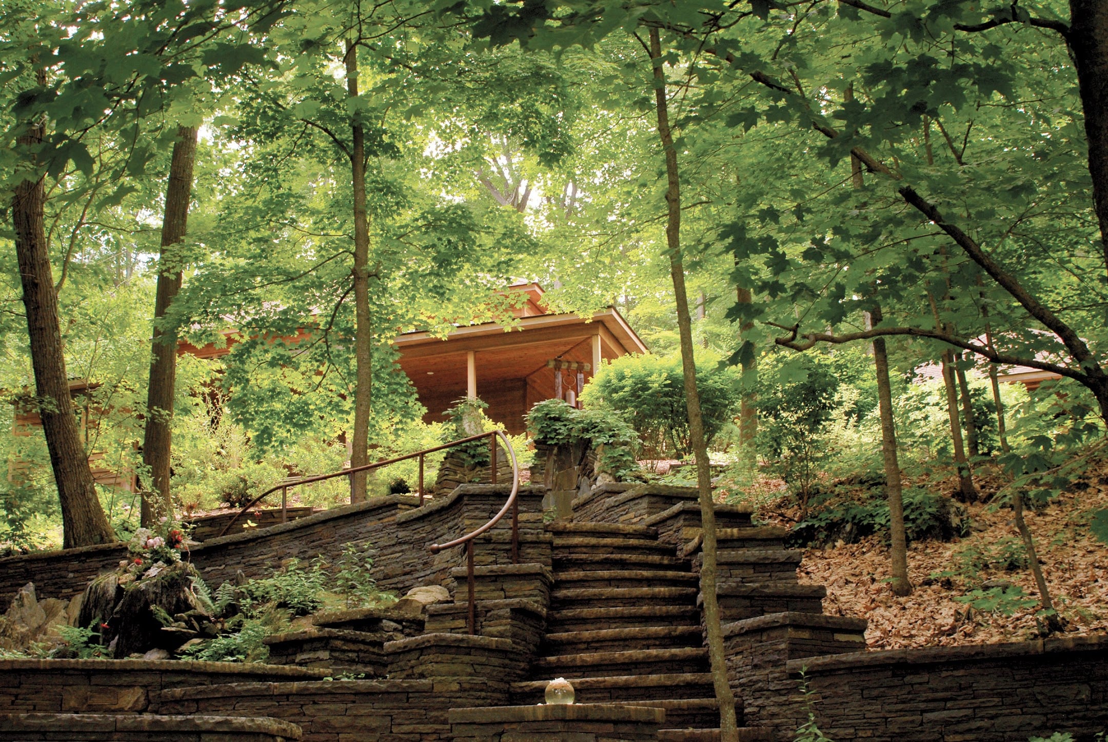 Stone steps with a curved handrail climb past terraced walls through leafy woods to a wooden pavilion, a peaceful retreat at Omega for meditaton, sound bath, and small workshop gatherings.