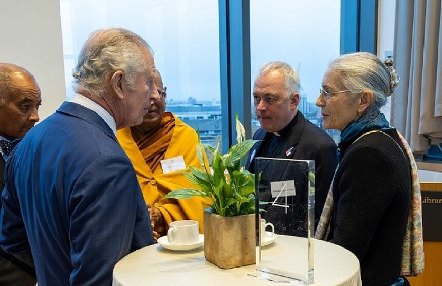 Five attendees at a round table during a formal event, chatting by coffee cups and a table 4 sign; a person in saffron robes and suited adults face a skyline, showing testing of leadership dialogue.
