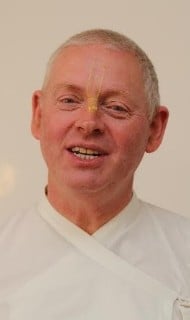 Close-up of a smiling person in white clothing with a thin gold line on the forehead, set against a plain light background; neutral portrait for an Articles feature on organized religion and faith.