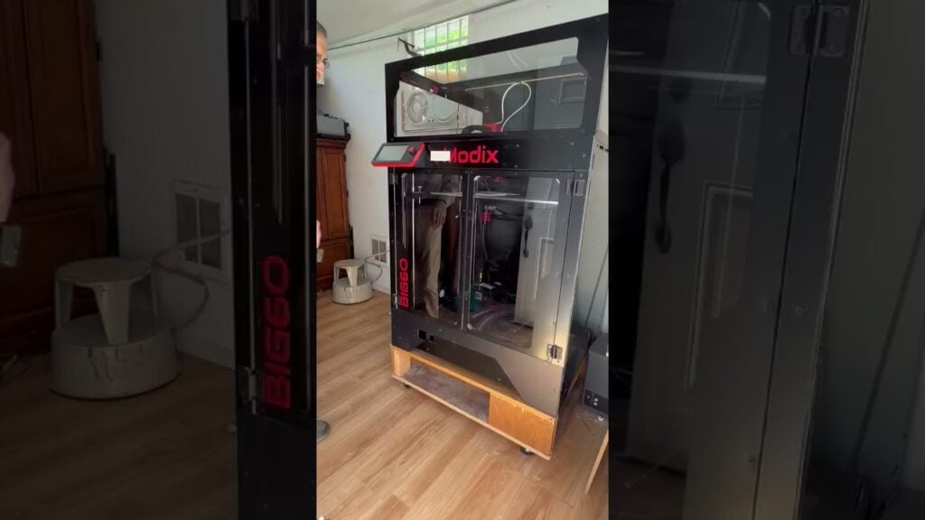 Large Modix BIG-60 enclosed 3D printer on a rolling wooden stand in a compact workshop with wood flooring and a cabinet; a person is partly visible as the machine is positioned for initial testing.