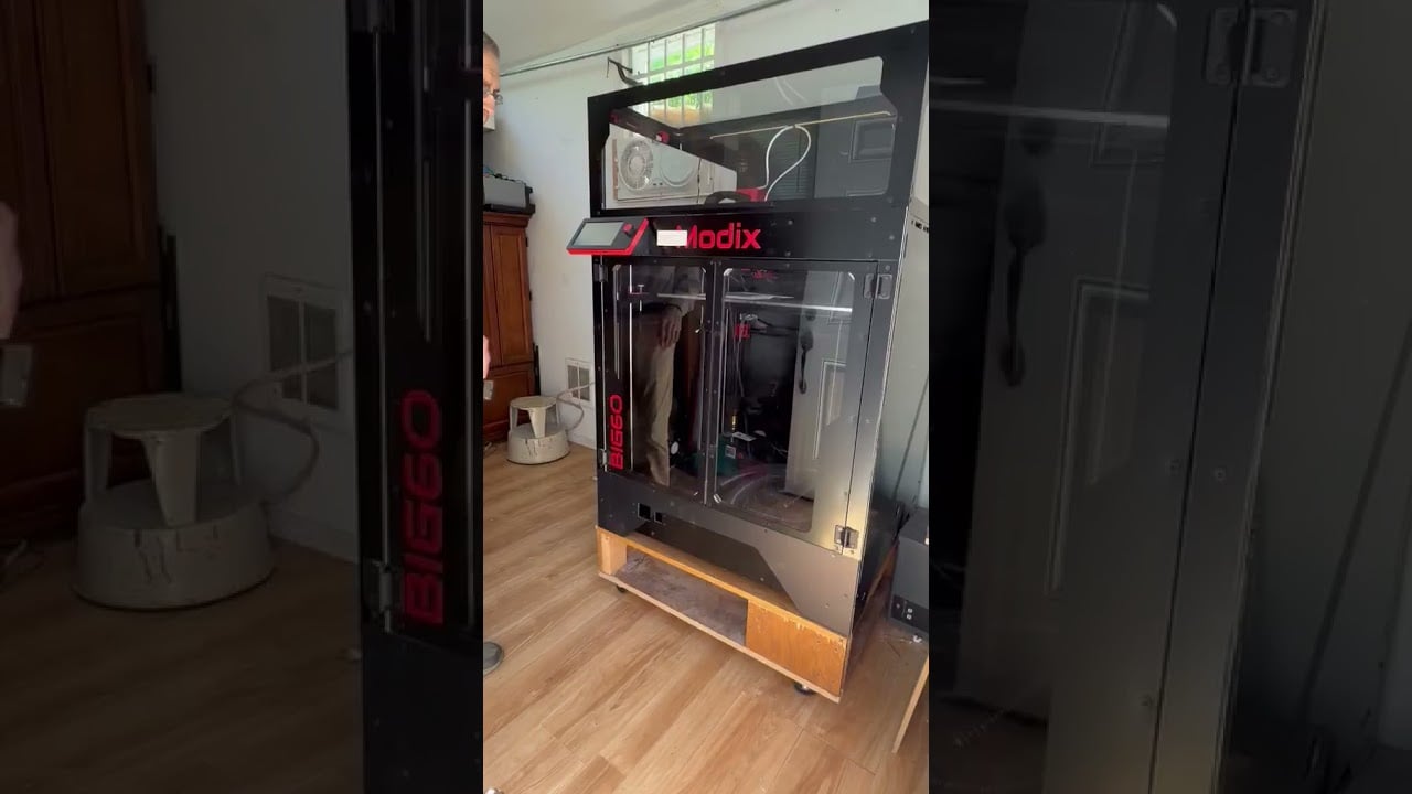 Large Modix BIG-60 enclosed 3D printer on a rolling wooden stand in a compact workshop with wood flooring and a cabinet; a person is partly visible as the machine is positioned for initial testing.