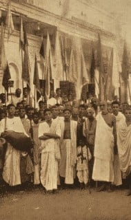 Sepia-toned crowd in traditional attire stands before a large building, with many flags and a hand drum visible—an image that evokes sankirtana, public kirtan processions, and welfare for all beings.
