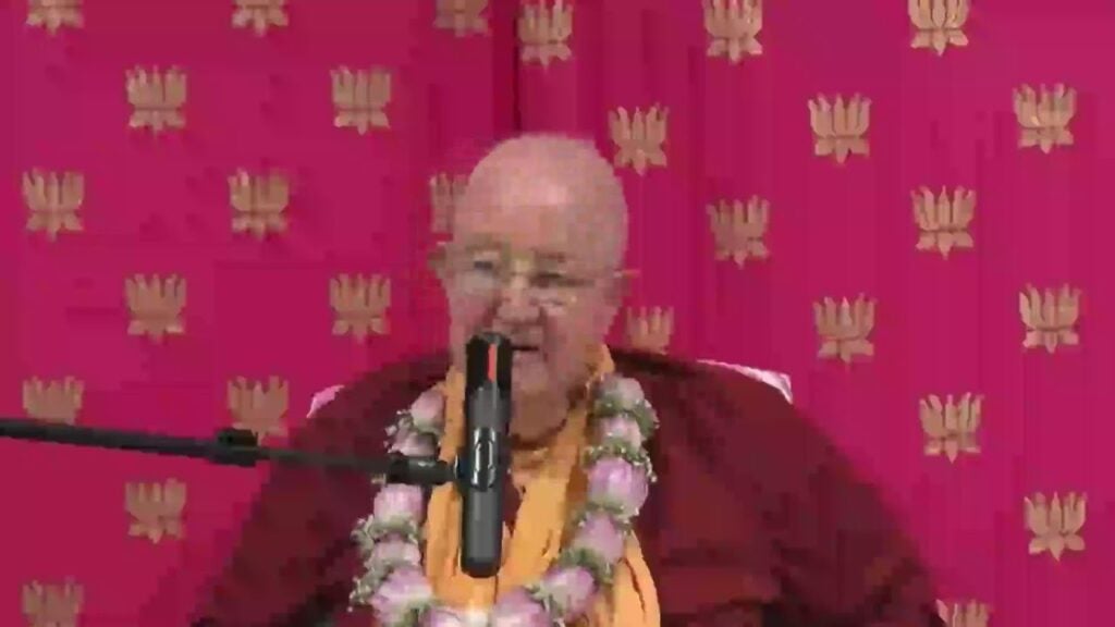 Speaker in maroon and saffron robes, wearing a pink flower garland, speaks into a microphone before a magenta backdrop with gold emblems; talk on envy and respect in Brambleton, testing.