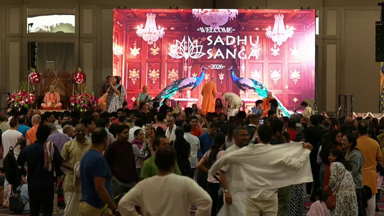 Day 1 of 2026 Sadhu Sanga: a crowded hall faces a stage where a welcome screen with chandeliers and peacocks glows behind saffron-robed monks, floral displays, and a seated figure; testing category.