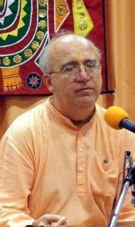 A speaker in saffron robes with glasses and a ceremonial forehead mark sits at a microphone before a colorful textile backdrop, illustrating our Articles discussion on faith, identity, and choice.