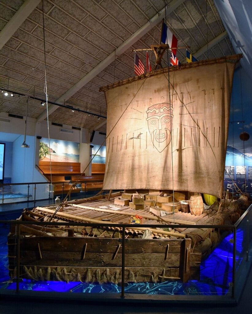 Exhibit of the Kon-Tiki balsa-wood raft, with lashed logs, cargo boxes, and a tall canvas sail painted with a tiki face - icon of Thor Heyerdahl's 1947 Pacific expedition and 5,000-mile crossing.