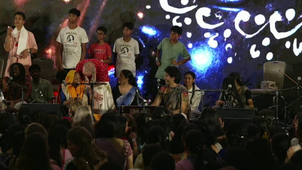 At 2026 Sadhu Sanga Day 3, performers lead a kirtan on stage with microphones and harmonium, children watch from behind, and a packed audience faces them beneath a swirling cosmic backdrop; testing.