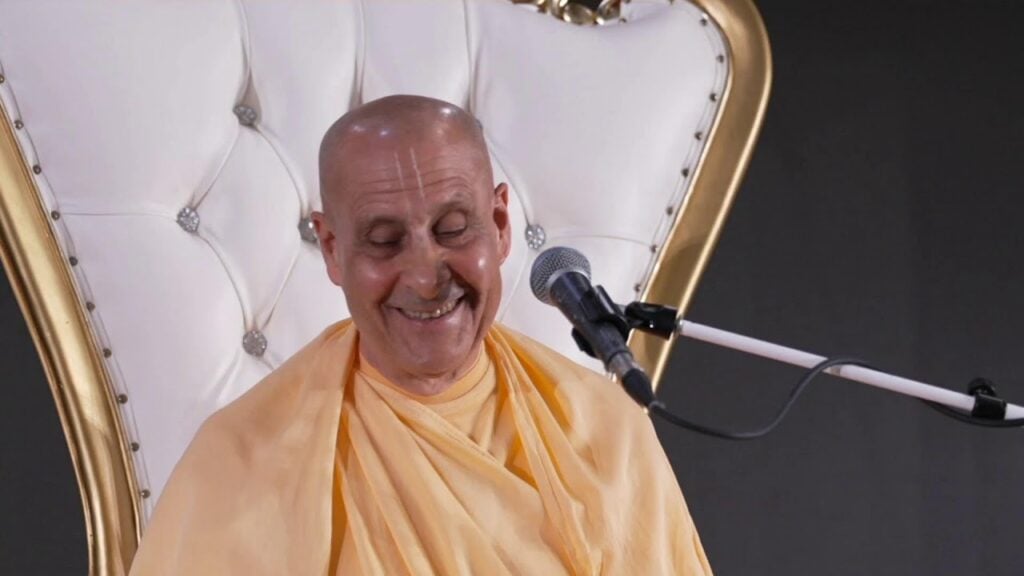 Close-up of a smiling, robed speaker seated on a white, gold-trimmed tufted chair beside a microphone during a public talk, eyes gently lowered and calm. testing.