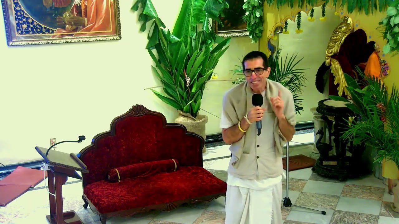 Devotional speaker in traditional attire holds a microphone in an ornate temple hall with settee, lush plants, and framed art, during a Los Angeles festival on Simple Living High Thinking; testing.