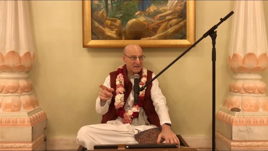 Garlanded teacher sits cross-legged on a temple dais, speaking into a microphone and pointing, with a framed painting and peach-white columns behind; morning class on SB 10.6.10; testing.