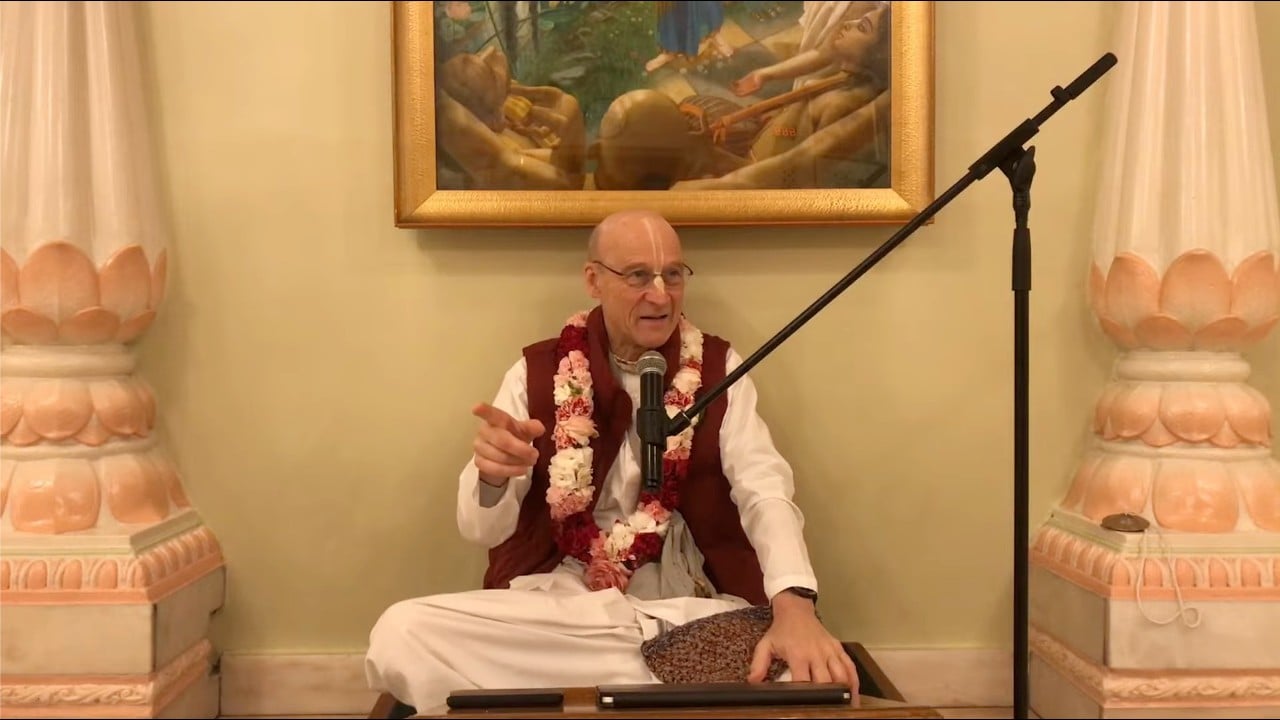 Garlanded teacher sits cross-legged on a temple dais, speaking into a microphone and pointing, with a framed painting and peach-white columns behind; morning class on SB 10.6.10; testing.