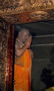Devotee in saffron robes prays in a dimly lit temple sanctum, hands joined before a dark statue, framed by ornate gold-toned carvings and a narrow doorframe.