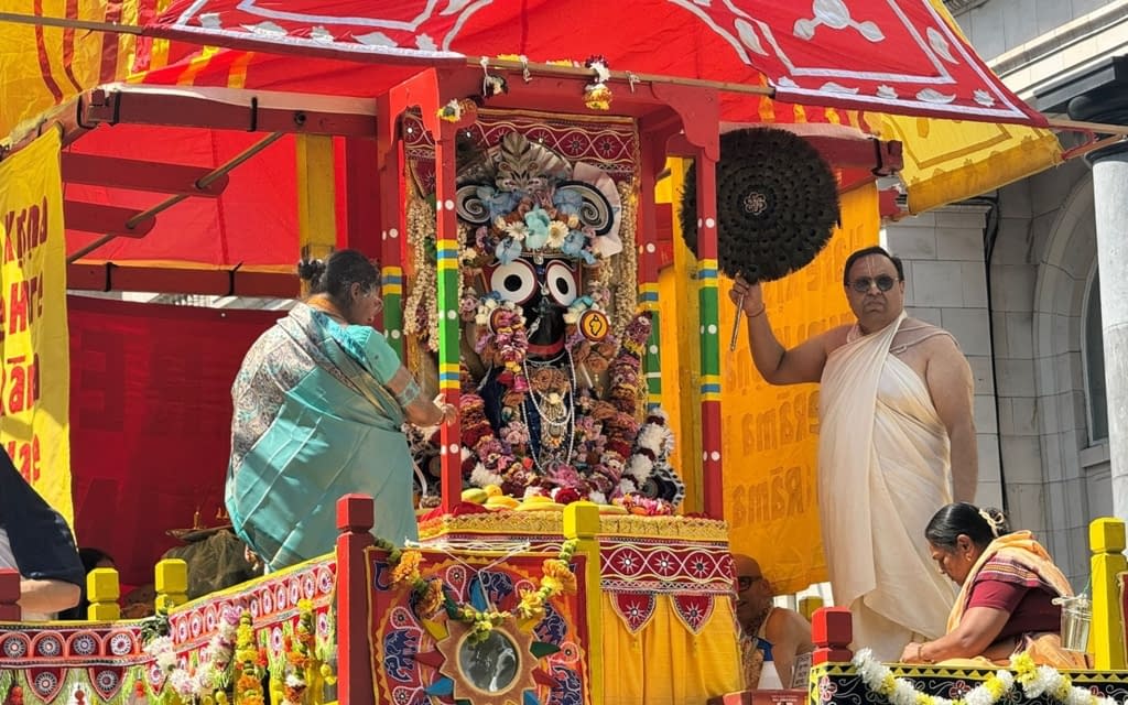 Brightly decorated Rath Yatra chariot carrying the Jagannath deity, garlanded with flowers, as priests and devotees perform rituals beneath a red-yellow canopy during London's Festival of Chariots.