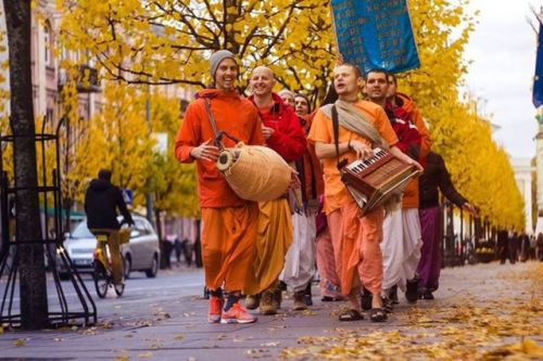 A group of people in orange robes walks along an autumn city street, smiling and singing while playing a hand drum and a small keyboard instrument beneath golden trees, a lively scene for testing.