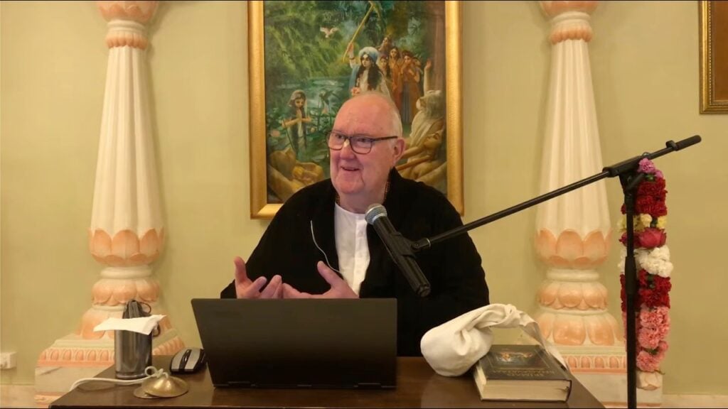 A speaker with glasses addresses a morning class from a desk with a laptop, mic, and scriptures, between ornate columns and a Krishna painting in a temple hall; event for SB 10.6.22–23 testing.