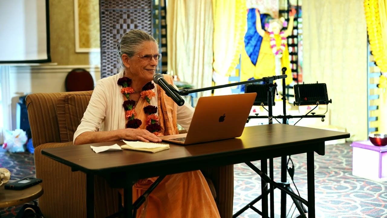 A speaker in saffron attire and a floral garland addresses a microphone from behind a laptop in a brightly decorated hall with an altar; scene from Vaishnava Sanga Festival 2025, Aug 1–4.