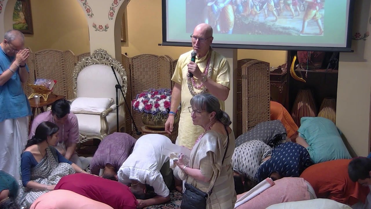 Krishna Katha temple gathering: a speaker with a microphone stands by an ornate chair as attendees bow in prayer; garlands, wicker screens, and a slide of Krishna art create a reverent scene.