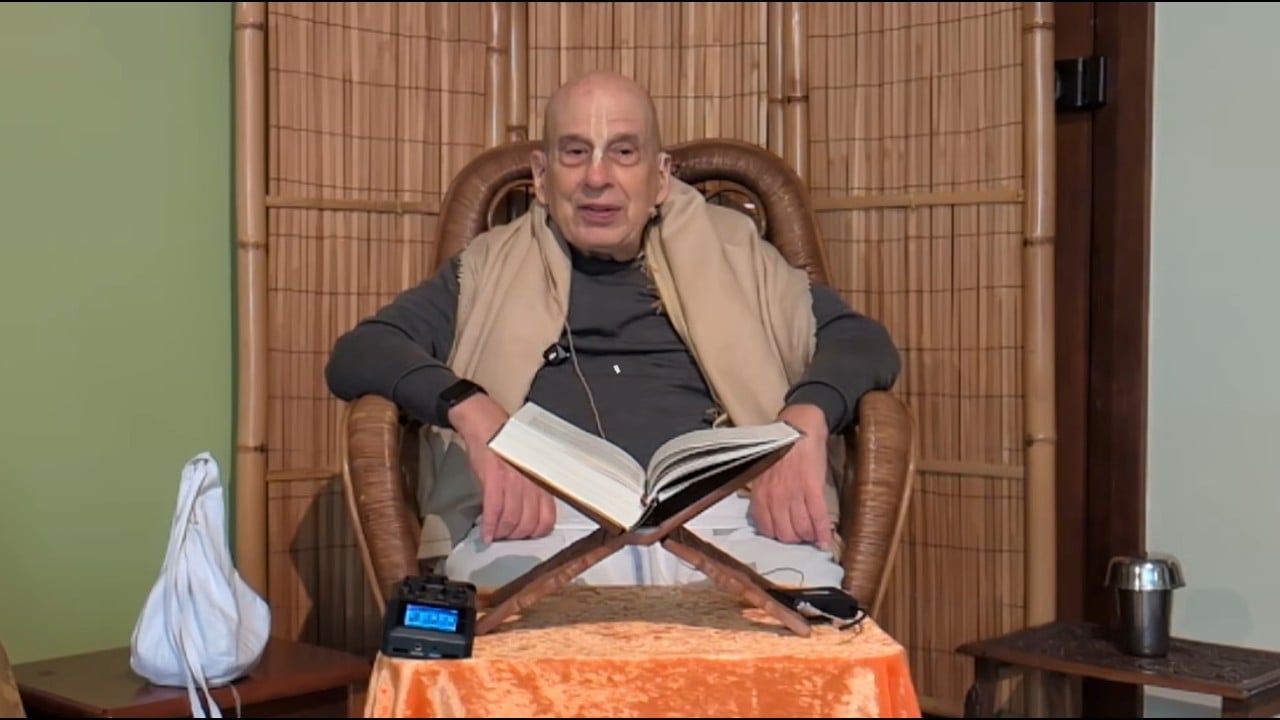 Speaker in a bamboo chair leads a Caitanya Caritamrta class, reading from an open book on a wooden stand; a mic recorder, bead bag, and metal cup sit nearby in a calm room. testing.
