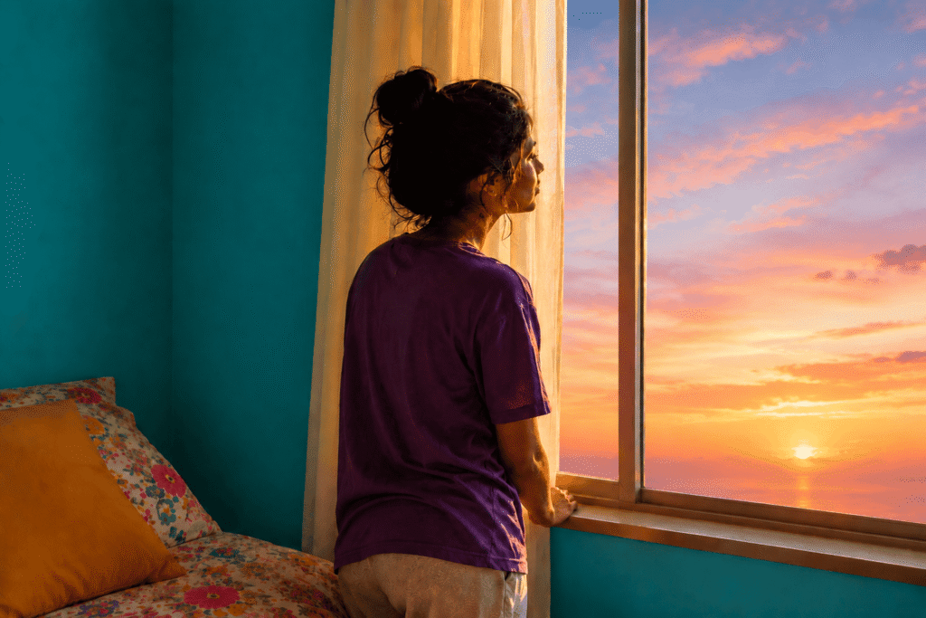 Person in a purple shirt stands by a bedroom window at golden hour, gazing at the sky; soft light, teal walls, and floral bedding evoke reflection, slow change, boundaries, healing, and personal growth.