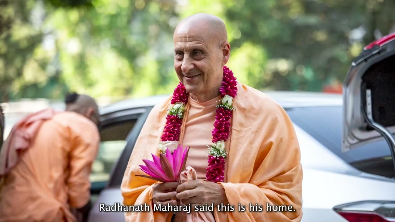 Smiling person in saffron robes and a flower garland holds a pink lotus beside a car, subtitles visible; still from a Govardhan Eco Village short film, testing category.