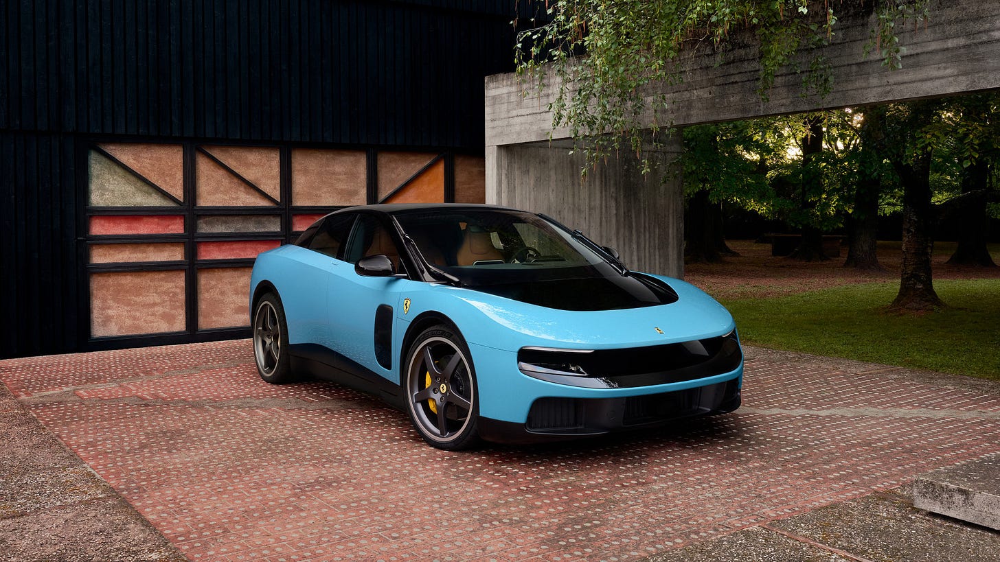Front three-quarter view of a sky-blue Ferrari coupe with black hood and roof, five-spoke wheels and yellow calipers, parked on a brick courtyard beside dark wood and concrete structures under trees.