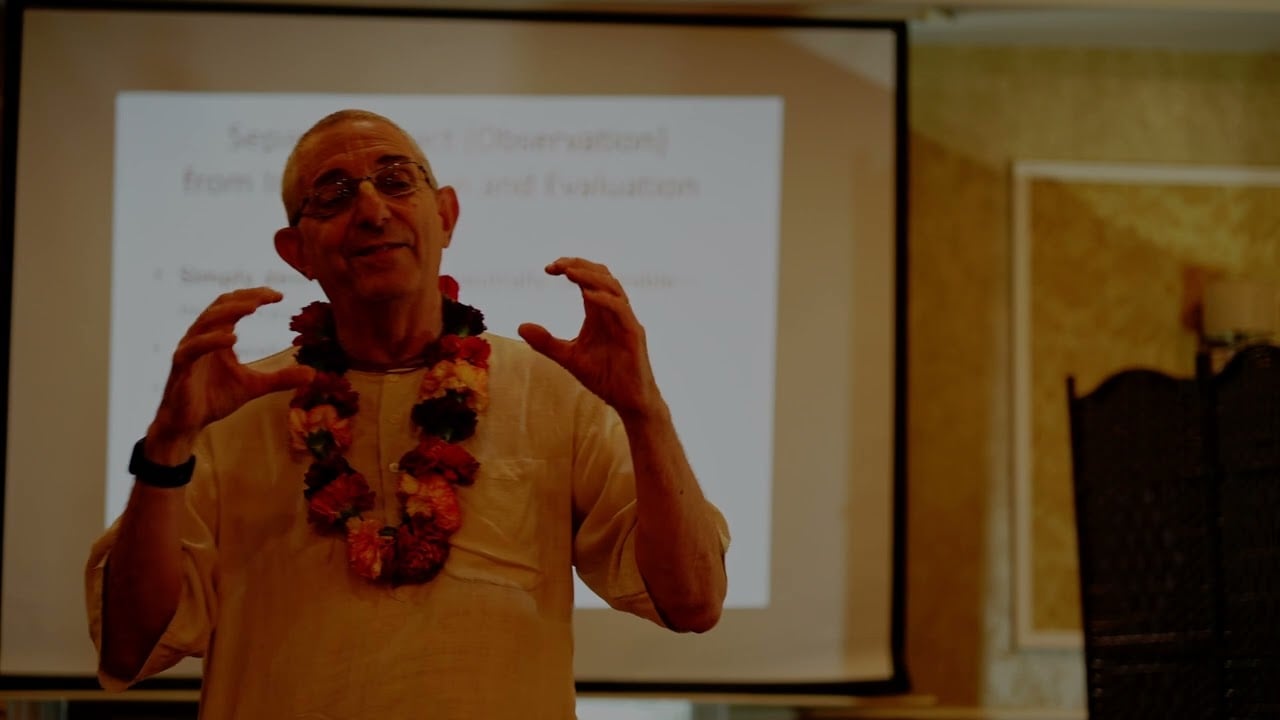 An older speaker in a cream kurta, glasses, and a flower garland gestures beside a projected slide in a warm-lit hall; image for a Vaishnava sanga 2025 lecture post, categorized under testing.
