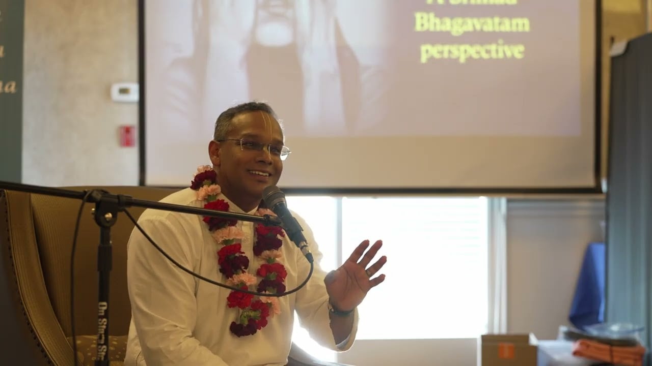 Speaker in white with a flower garland addresses the audience via microphone, gesturing as a slide reads 'Bhagavatam perspective'; talk on 11 causes of dejection, part 2, for testing content.