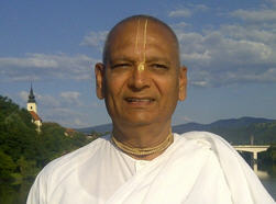 Portrait of a smiling man in white traditional clothing with a vertical forehead mark, standing outdoors by hills, river, and a tower under blue sky; for a senior devotee glorification post, testing.