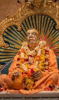 Garlanded statue of a Vaishnava guru in saffron robes, seated on an ornate throne with a radiant blue-gold halo backdrop, tilaka markings, and marigolds, in a bhakti temple setting.