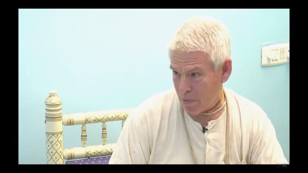 Video still of an older man with short white hair, wearing a cream robe and necklace, seated by a carved bed against a pale blue wall, looking right while speaking for an SB 4.18.19 testing post.