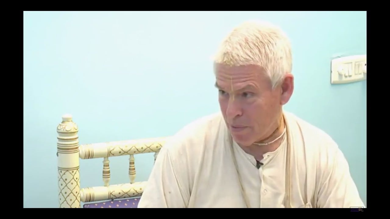 Video still of an older man with short white hair, wearing a cream robe and necklace, seated by a carved bed against a pale blue wall, looking right while speaking for an SB 4.18.19 testing post.