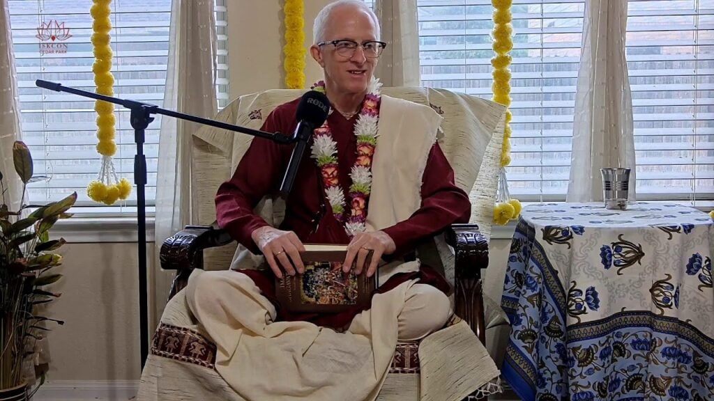 Speaker in maroon kurta and cream shawl sits cross-legged with a mic, garland, and scripture, beside a patterned table and window garlands at ISKCON Cedar Park; Srimad Bhagavatam class SB 2.5.11.