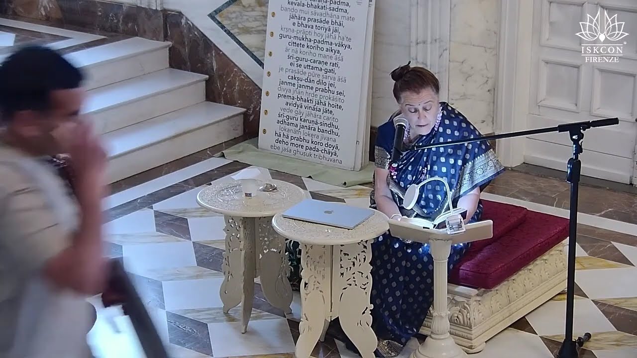 A speaker in a blue sari reads from a text at a low lectern with mic in an ISKCON Firenze hall; carved white tables, a laptop, and a verse board signal a retreat session on S.B. 5.18.9; testing