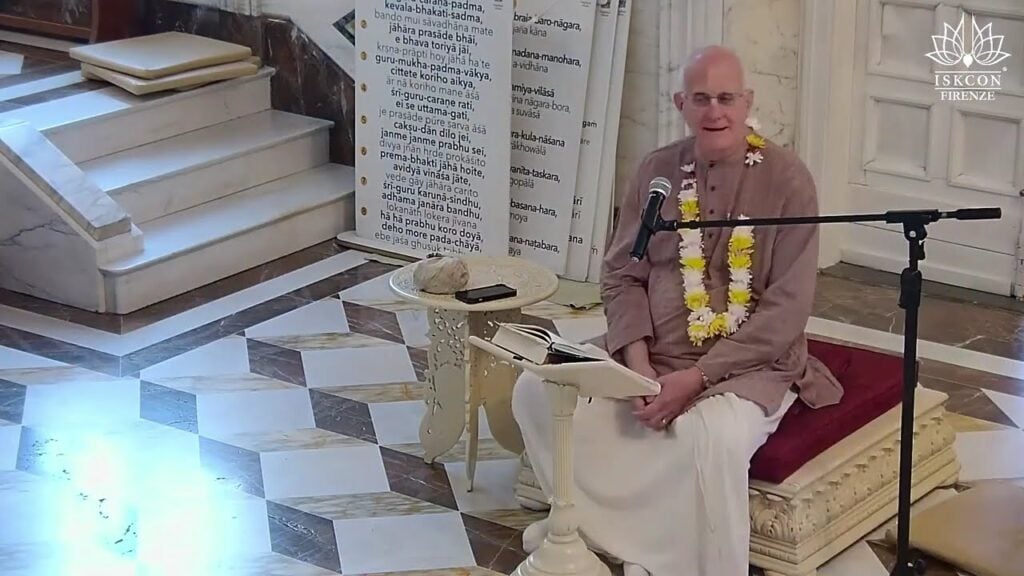 Speaker on a carved platform, wearing a flower garland, with mic and open scripture in a marble-floored ISKCON Firenze hall; verse banners hint at a Srimad Bhagavatam 1.2.6 testing livestream.