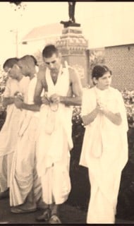 Archival sepia photo of Hare Krishna devotees in white robes walking outdoors, fingers on japa beads, echoing the early roots of Krishna Lunch and its campus service.