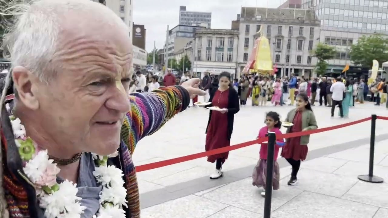 Nottingham Rathayatra 2026 in the city centre: a garlanded man points toward a tall yellow chariot and a festival crowd, while two children walk past with plates behind a red barrier. SEO testing.