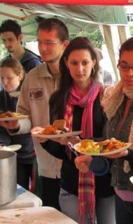 Queue of people under a green tent at an outdoor food stall, holding plates filled with salad, bread, and hot dishes while passing a large metal pot on a serving table in a buffet-style line.