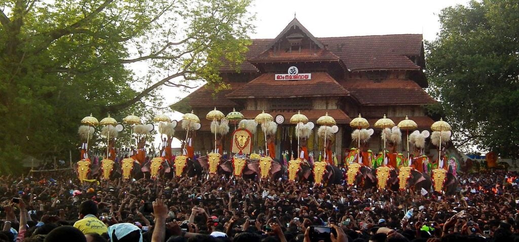 Caparisoned elephants line up with golden headpieces and silk parasols before a crowd at a Kerala temple festival, framed by large trees and a tiled-roof shrine—a classic Thrissur Pooram scene.