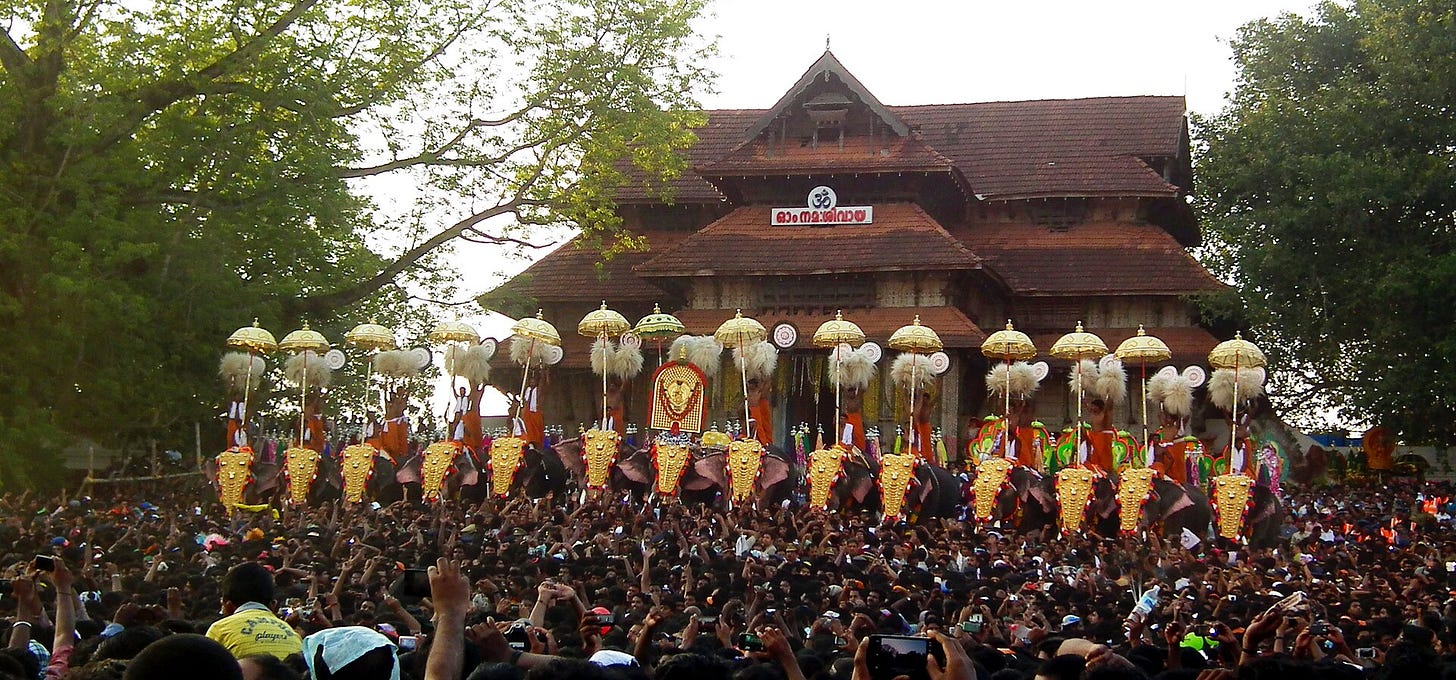Caparisoned elephants line up with golden headpieces and silk parasols before a crowd at a Kerala temple festival, framed by large trees and a tiled-roof shrine—a classic Thrissur Pooram scene.