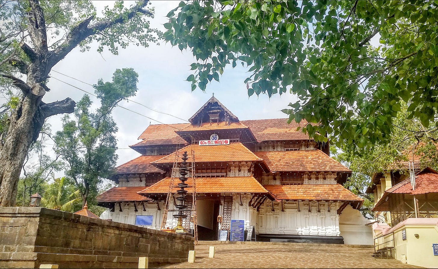 Historic Kerala temple with tiered red-tiled roofs and white carved walls, framed by banyan and peepal leaves; a deepasthambham oil lamp stands near the central entrance in a stone courtyard.