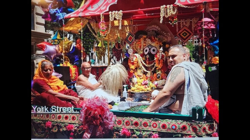 Vibrant Ratha Yatra chariot in NYC carrying the flower-garlanded Jagannath deity, with fruit offerings, incense, and devotees in traditional attire beneath a red canopy and balloons.